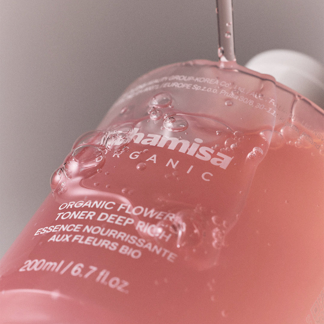 Flacon rose de lotion tonique WHAMISA Organic Flowers Deep Rich, avec un filet de liquide qui coule sur l’étiquette