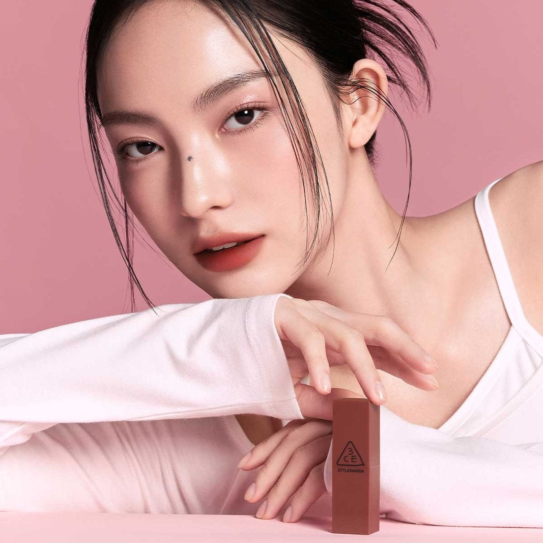 Décris l'image en français : une jeune femme maquillée, tenant le 3CE Mood Recipe Lip Color de 3CE sur fond rose.
