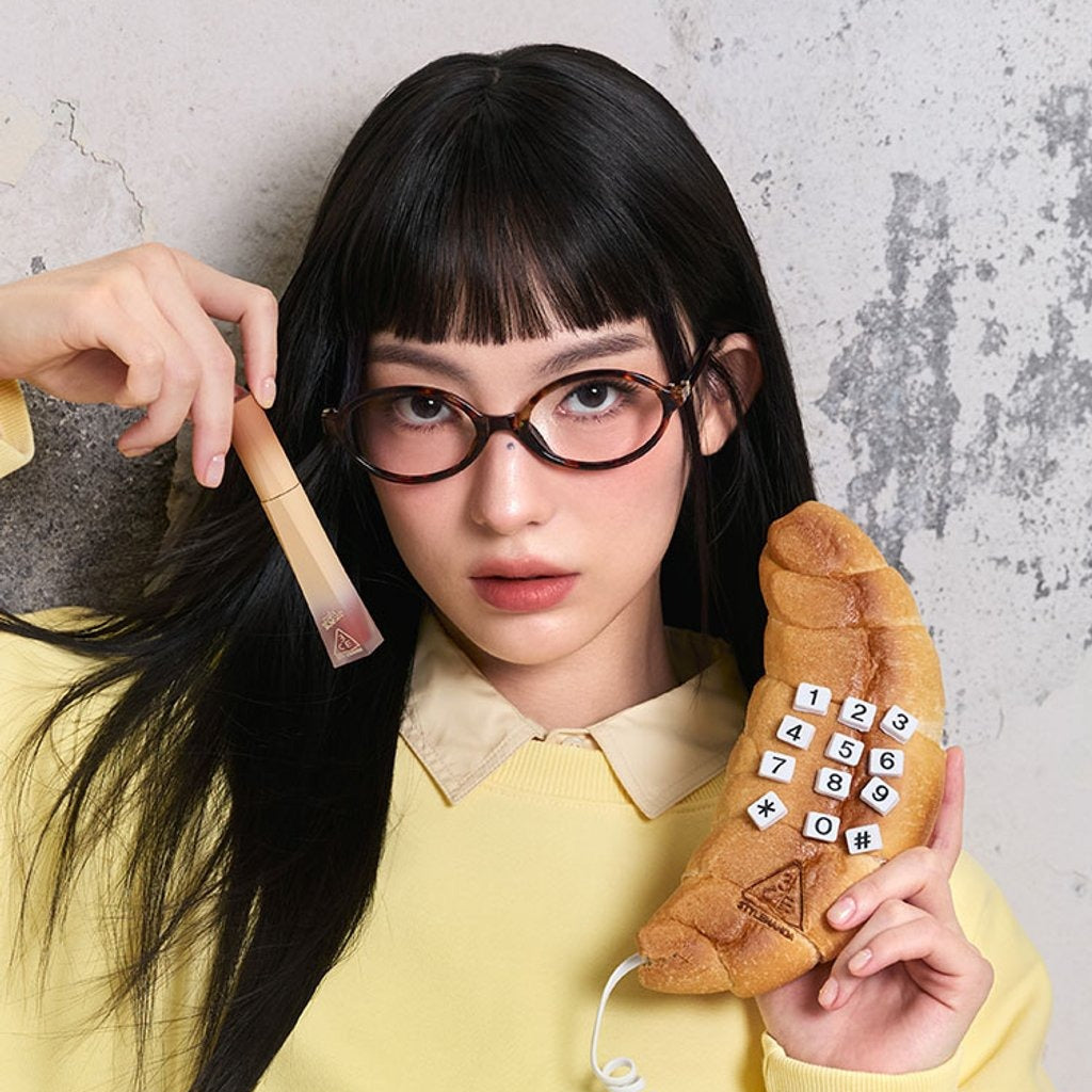 Décris l'image en français : 3CE COLOR BAKER VELVET LIP TINT PLUSH, marque 3CE, maquillage sur fond mural avec femme aux lunettes.