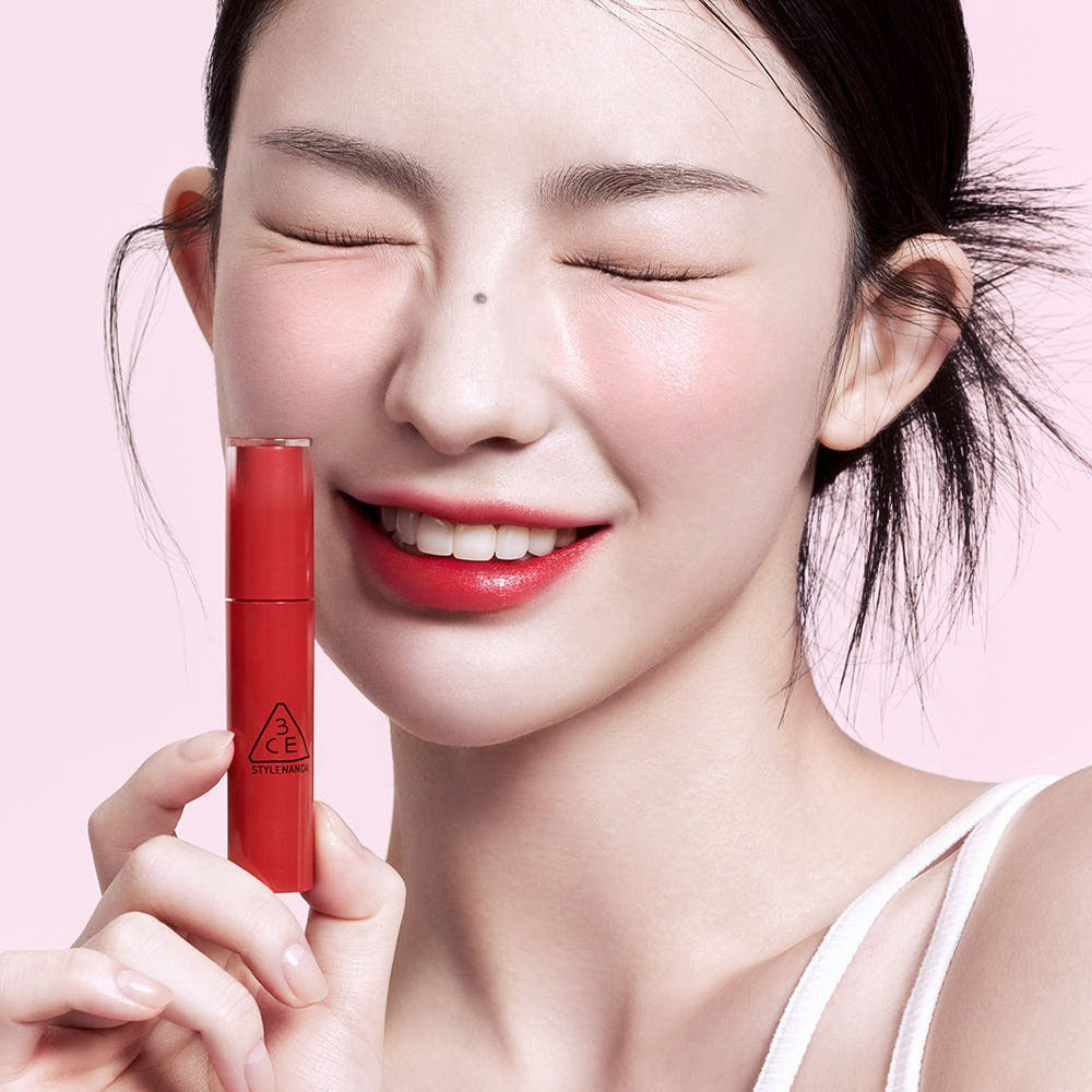 Modèle souriant tenant le 3CE Lazy Pop Lip Stain, mettant en valeur ses lèvres parfaitement colorées.