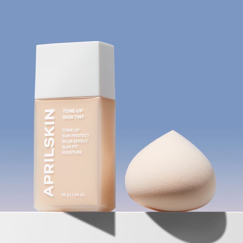 APRILSKIN Tone-Up Skin Tint d’APRIL SKIN, flacon et éponge maquillage sur fond bleu ciel, image produit.