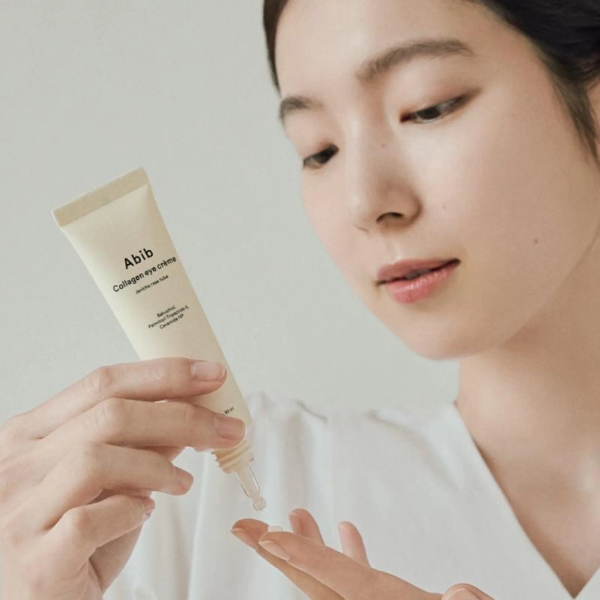 Décris l'image en français : ABIB Collagen Eye Cream Jericho Rose Tube 30ml d’Abib, main applique crème sur le visage, rose.
