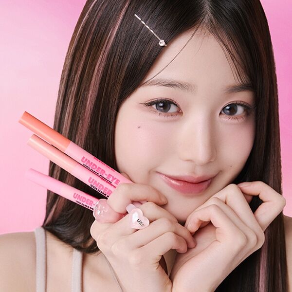 AMUSE Under Eye Volume Pencil de la marque AMUSE, sur fond rose, maquillage visage et regard, main tenant le produit.
