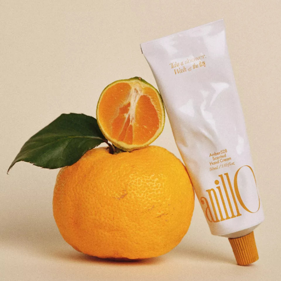 ANILLO Amber528 Scented Hand Cream 50ml, tube à côté d’orange et feuilles, fond beige, ambiance cosmétique artisanale.