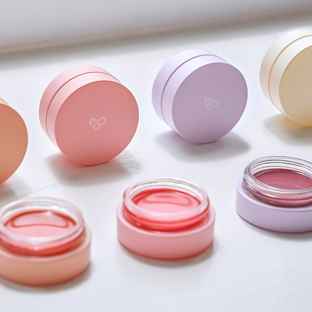 AOU Glowy Tint Balm de la marque AOU, pots et sticks colorés, ambiance maquillage sur fond blanc.