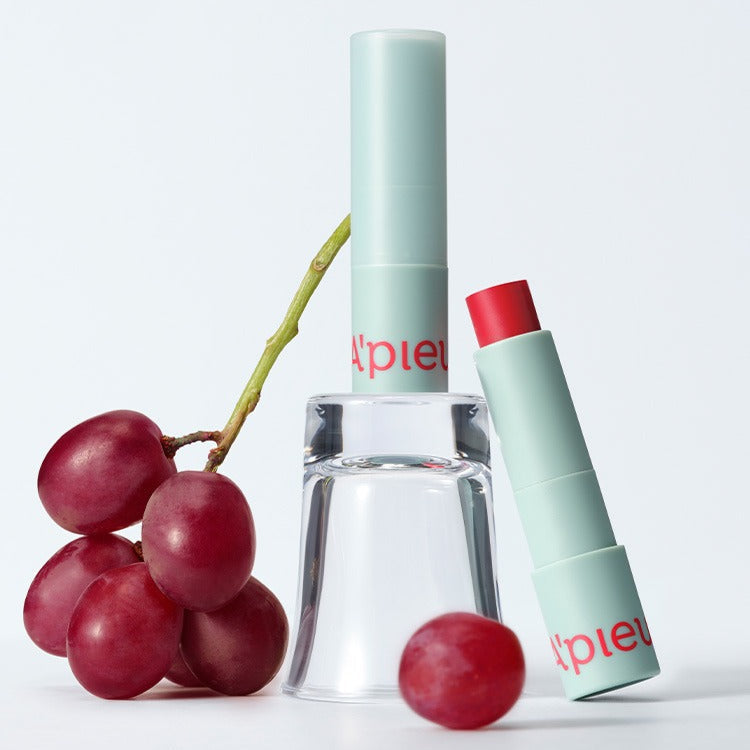 A'PIEU Juicy Pang Care Lip Balm, flacon vert sur fond blanc avec raisins rouges, marque Apieu en contexte beauté