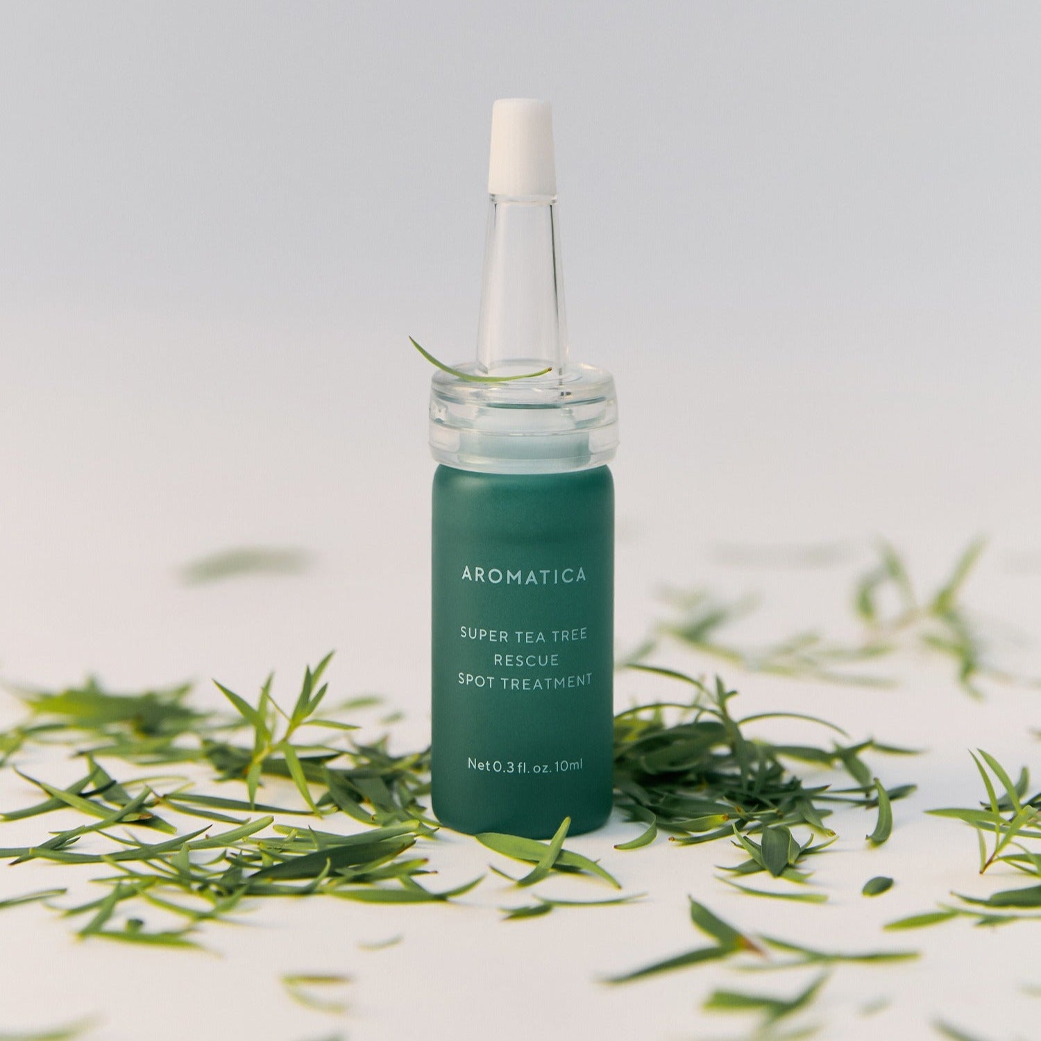 AROMATICA Super Tea Tree Rescue Spot Treatment 10ML posé sur un fond blanc avec des feuilles de thé autour, flacon de 10 ml visible.
