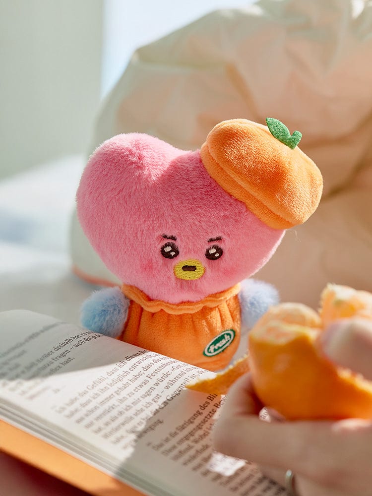 BT21 TATA BABY PLUSH ORANGE PARTY, peluche orange de V de BTS, posée sur un livre, décor bébé à la lumière douce.