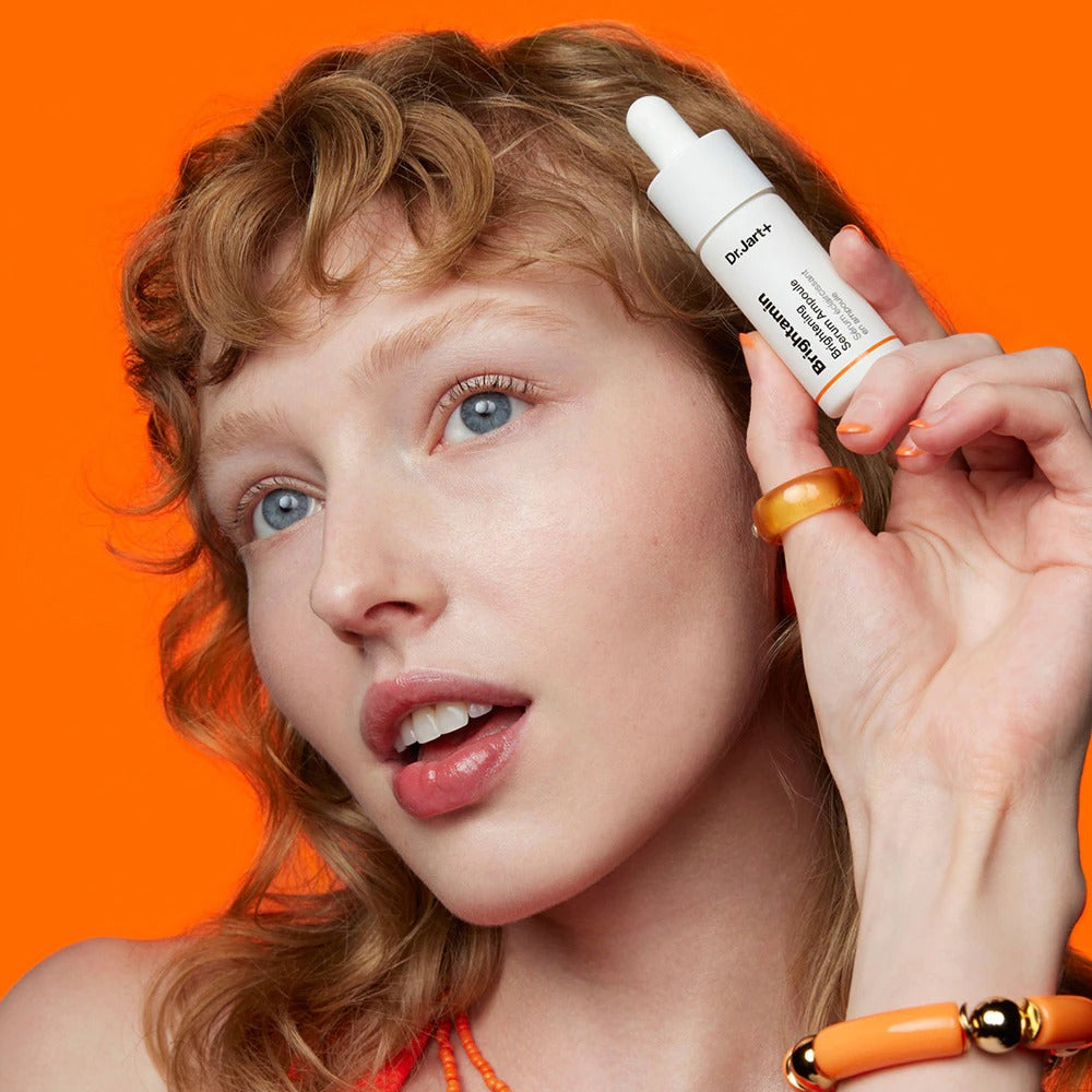 Femme posant avec le sérum DR.JART+ Brightamine Brightening Serum Ampoule sur un fond orange vibrant.