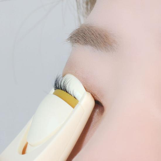 FILLIMILLI USB Heating Eyelash Curler : lisseur chauffant appliqué sur les cils, main tenant l’outil, fond clair.