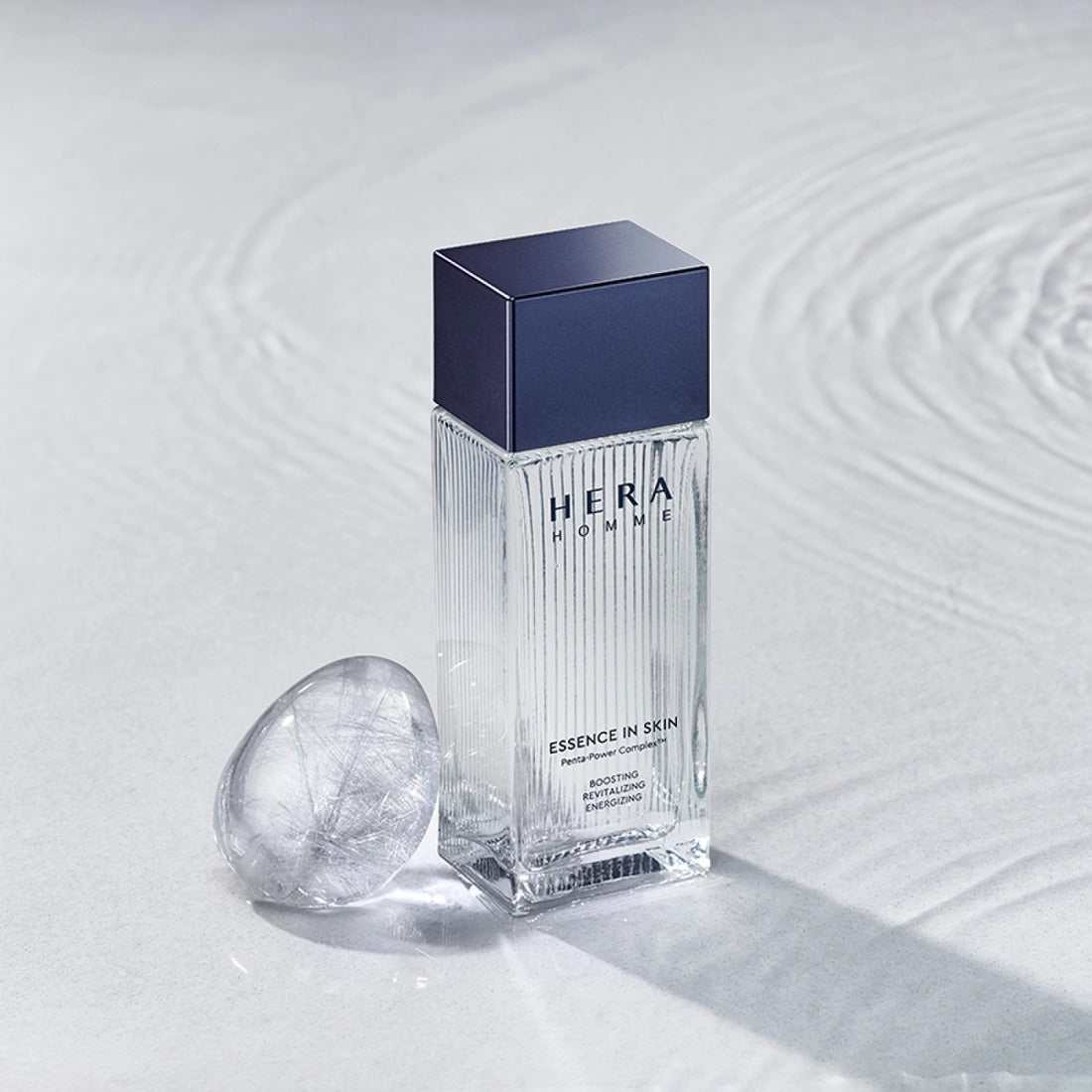 Décris l'image en mentionnant HERA Homme Essence In Skin 125ml, flacon HERA bleu sur fond blanc texture peau, contexte soin.