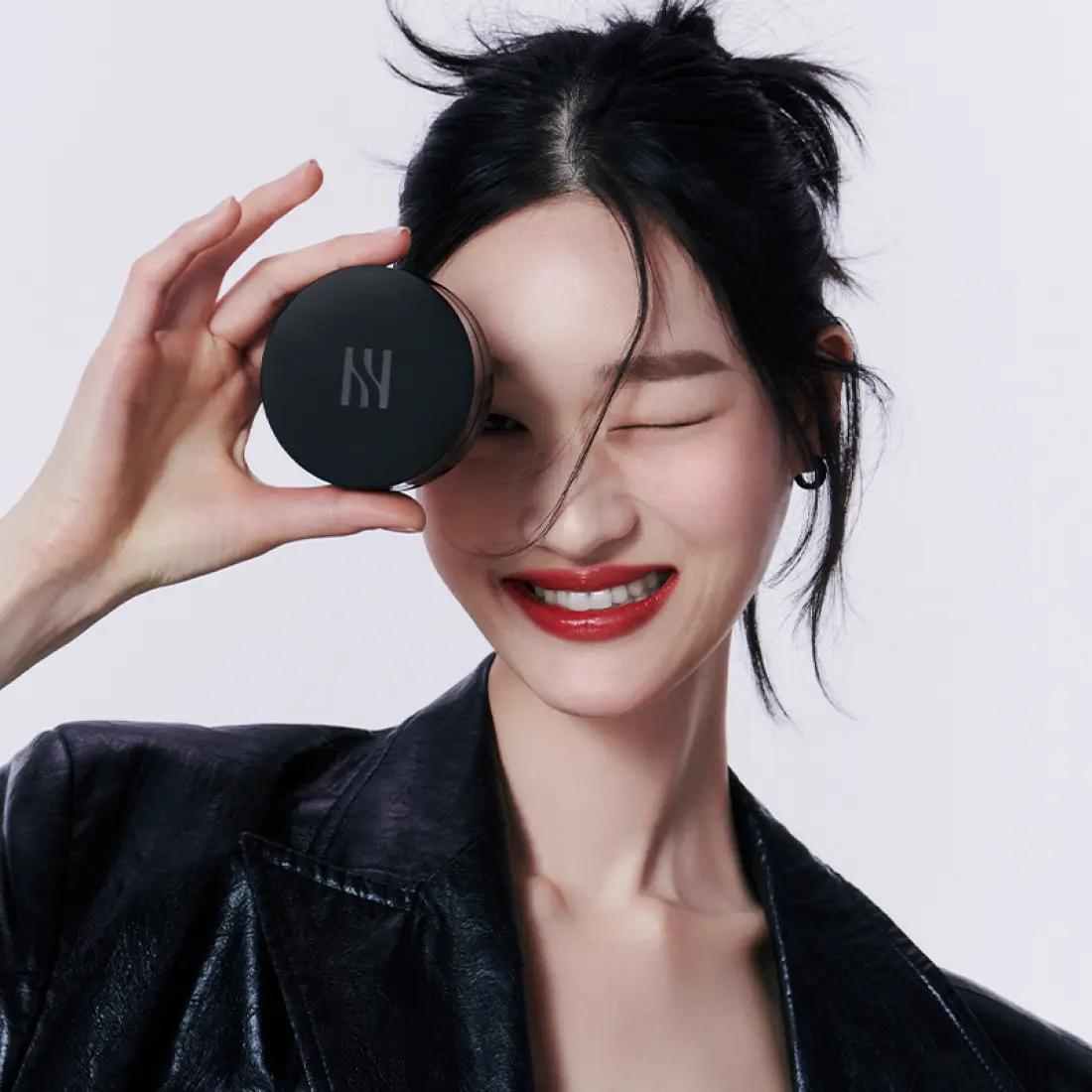 HERA Black Cushion Foundation SPF34 PA++ 15g de HERA, pot noir tenu près du visage sur fond clair.