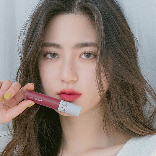 Portrait maquillage: ROMAND Zero Velvet Tint (#02 to #17) sur lèvres, marque ROMAND, fond clair, main tenant le produit.