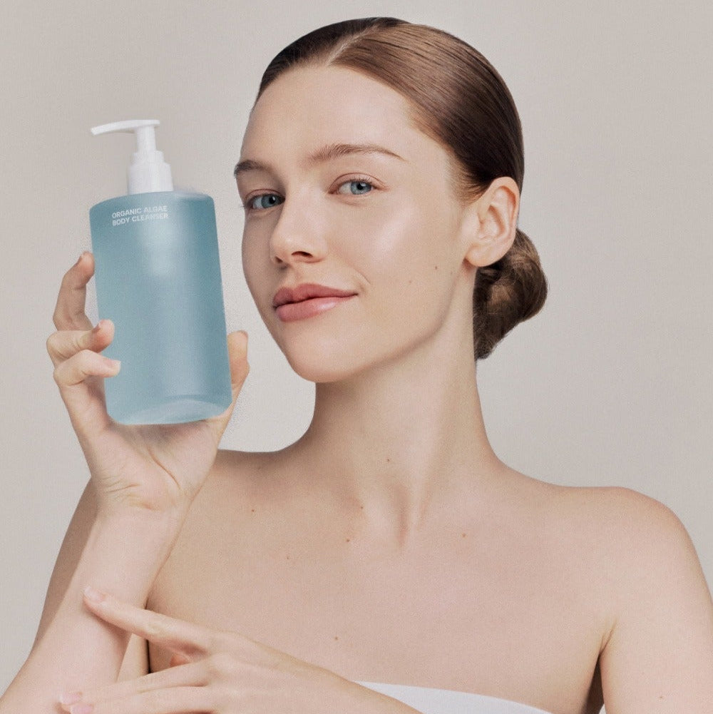 WHAMISA Organic Algae Fermented Body Cleanser 490ml, marque WHAMISA, femme montrant le flacon bleu en salle de bain.