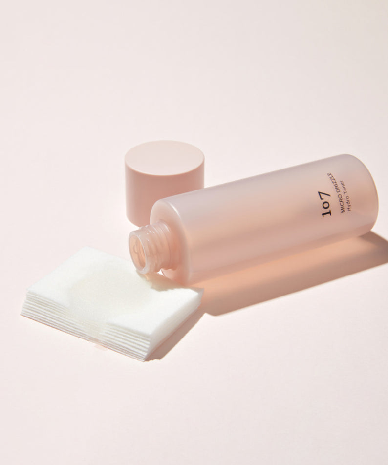 Bouteille de toner 107 Micro Drizzle Hydro Toner 155ml posée sur une surface rose avec des cotons.