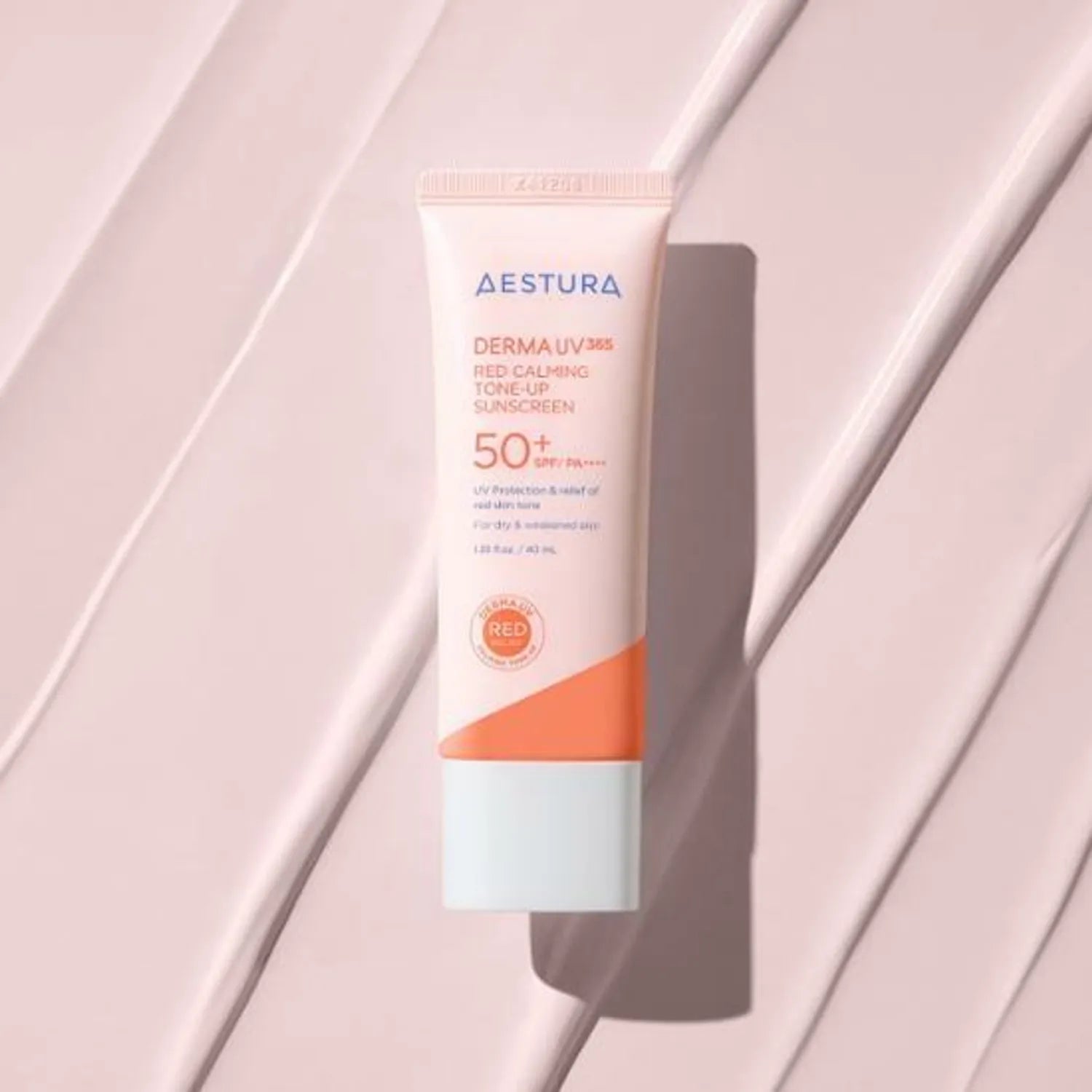 Décris l'image en français : AESTURA Derma UV365 Red Calming Tone-Up Sunscreen 40ml, tube AESTURA sur tissu rose