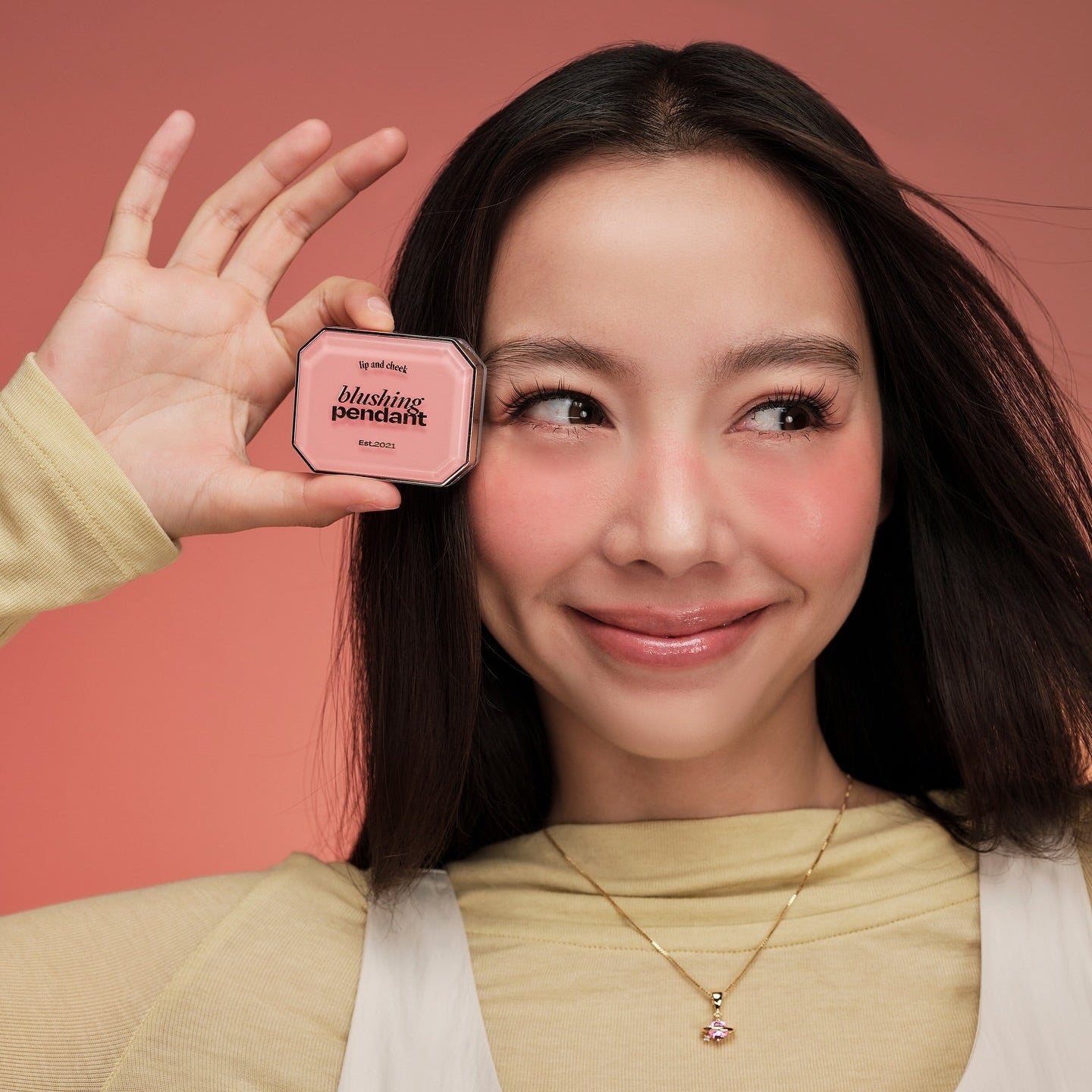 Décris l’image en français : ALTERNATIVE STEREO Blushing Pendant Lip & Cheek, femme sur fond rose tenant le produit