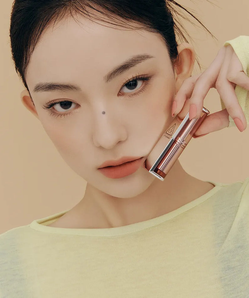 3CE Blur Matte Lipstick de 3CE, maquillage lèvres nude, modèle tenant un rouge à lèvres dans un décor beige