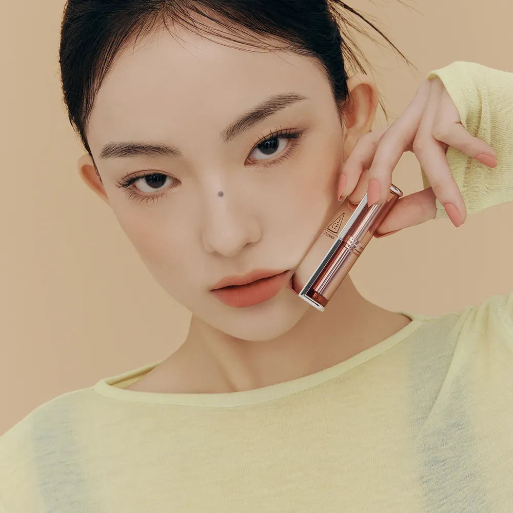 3CE Blur Matte Lipstick de 3CE, maquillage lèvres nude, modèle tenant un rouge à lèvres dans un décor beige