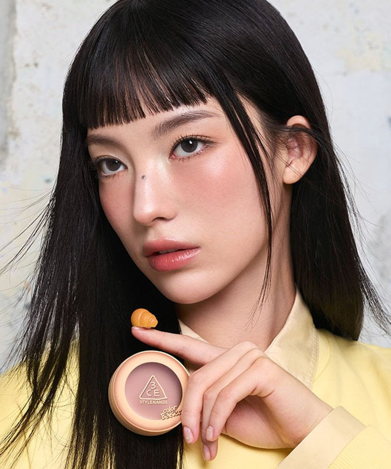 Décris l'image en français : 3CE Color Baker Face Blush, marque 3CE, maquillage sur le visage, jeune femme, pot rose maquillé.