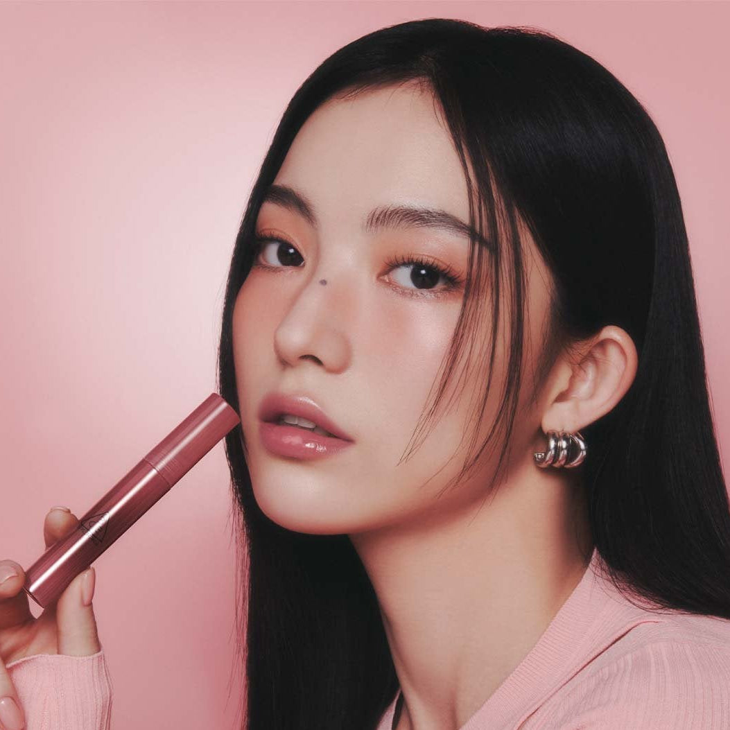 3CE Drop Glow Gel 10 Shades, la marque 3CE sur lèvres hydratées; femme maquillée tenant un baume, fond rose