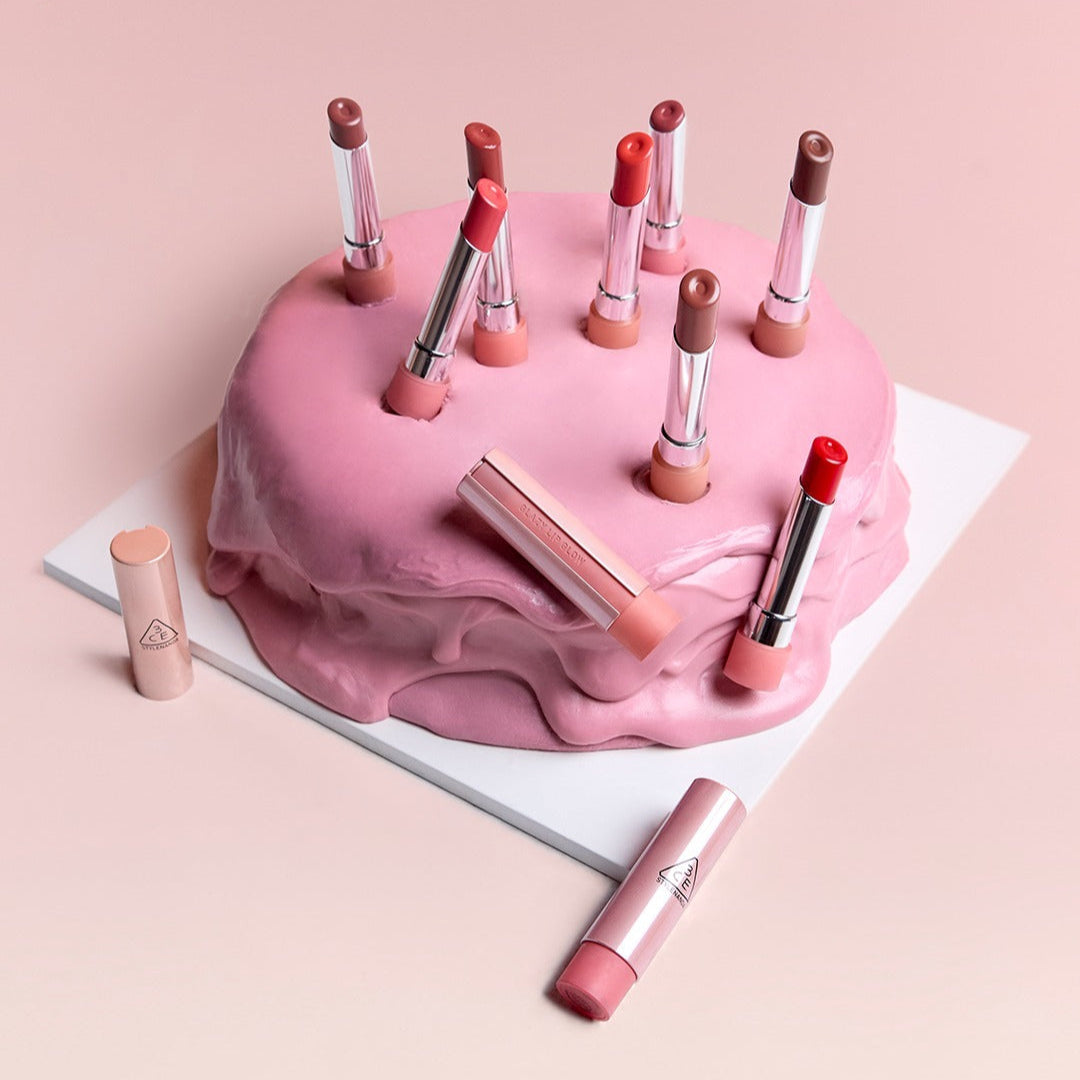 3CE Glaze Lip Glow 3.2g, baume brillant sur fond rose avec tubes et embouts maquillage 3CE, ambiance glamour.
