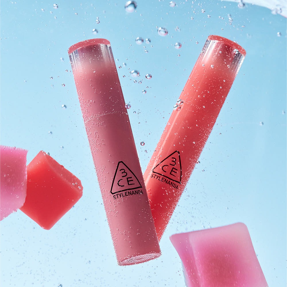 3CE Lazy Pop Lip Stain de StyleNANDA, tubes rouge rosé et bouchons en contexte éclaboussures d’eau sur fond bleu.