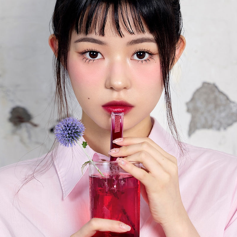 3CE Misty Lip Bare, femme aux lèvres rouges tenant le produit devant un verre rouge, décor floral en fond.