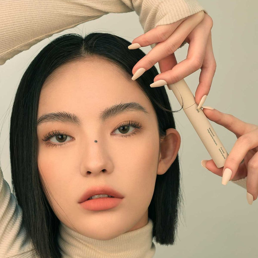 3CE Waterproof Mega Volume Mascara, flacon beige tenu près des cils d’une femme en gros plan (contexte maquillage).