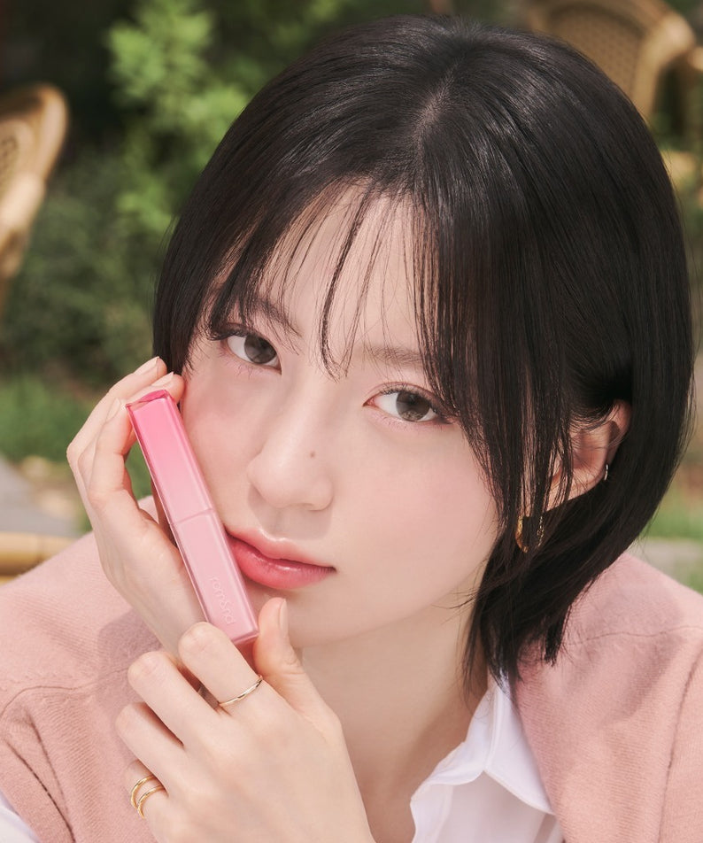 Portrait maquillage : ROMAND Sheer Tinted Stick de ROMAND, jeune femme en extérieur, main tenant le stick rose.