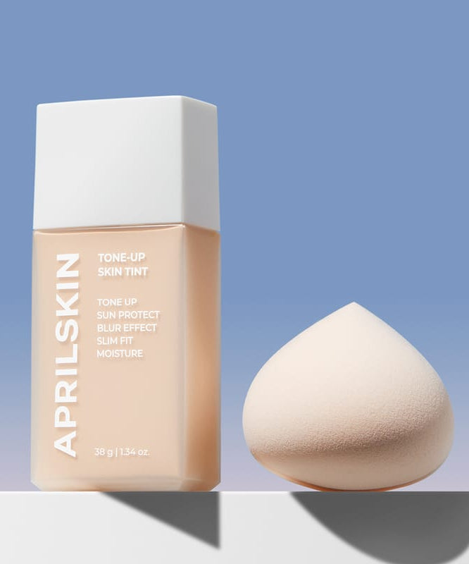APRILSKIN Tone-Up Skin Tint d’APRIL SKIN, flacon et éponge maquillage sur fond bleu ciel, image produit.