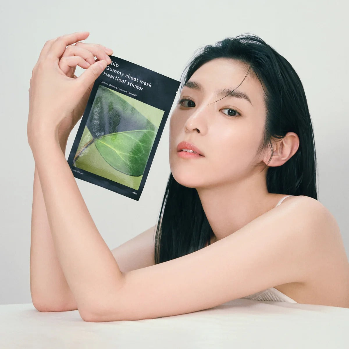 Femme appliquant ABIB Gummy Sheet Mask Heartleaf Sticker : masque feuille cœurleaf, produit ABIB sur fond clair.