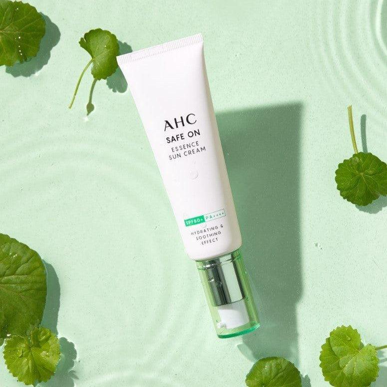 Décris l'image en français: AHC Safe On Essence Sunscreen 50ml, tube crème solaire AHC vert, feuilles, fond clair.