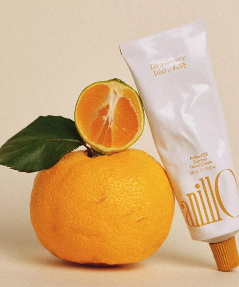 ANILLO Amber528 Scented Hand Cream 50ml, tube à côté d’orange et feuilles, fond beige, ambiance cosmétique artisanale.