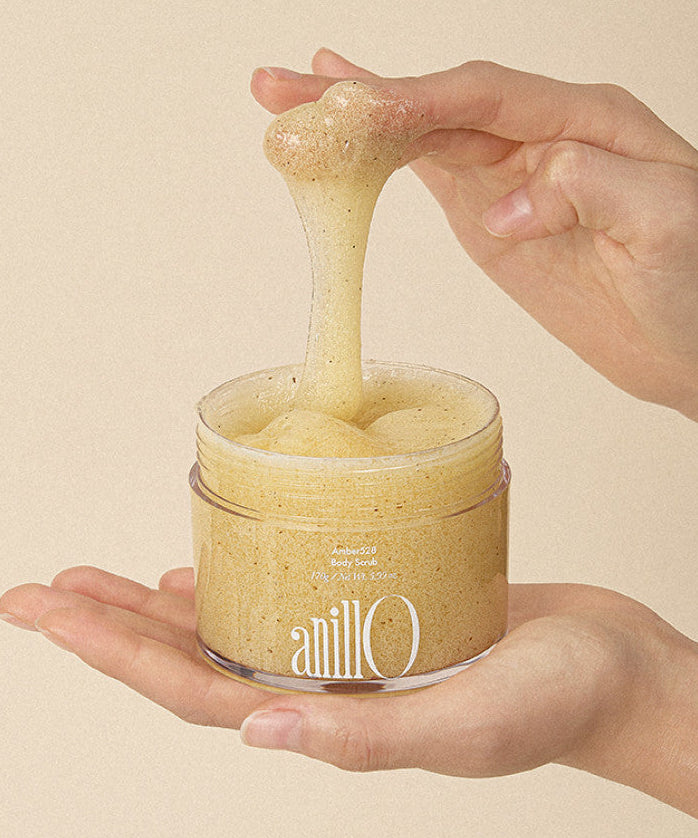 ANILLO Amber528 Body Scrub 170g en pot, mains tenant le gommage à grains sur fond beige.