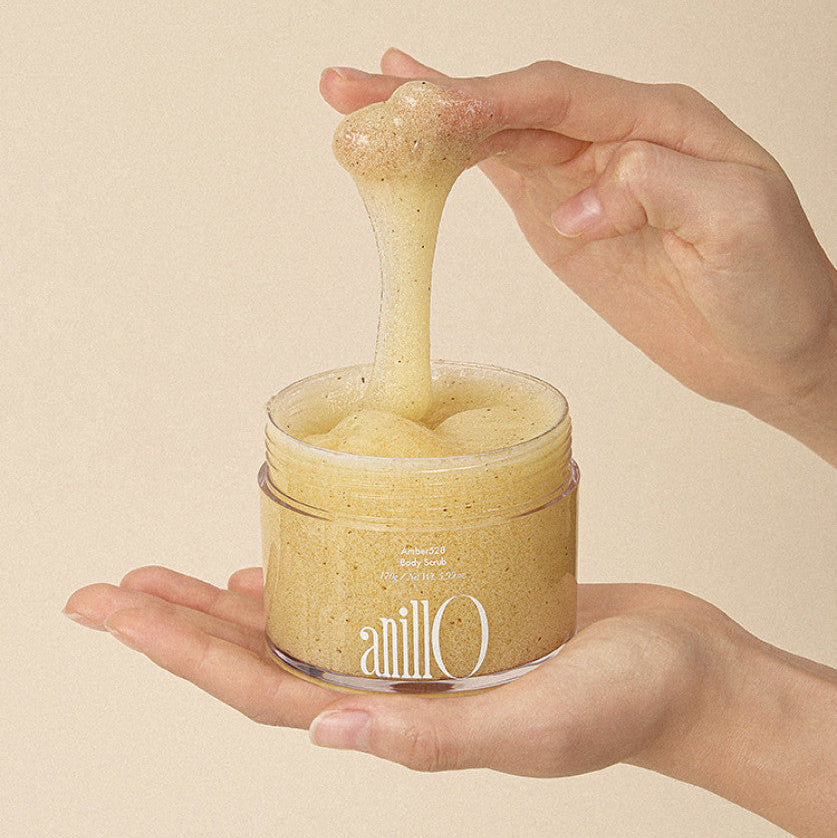 ANILLO Amber528 Body Scrub 170g en pot, mains tenant le gommage à grains sur fond beige.