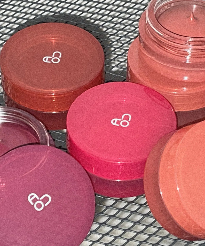 AOU Floofy Matte Balm de la marque AOU, baumes mats colorés en pots sur filet métallique, ambiance maquillage