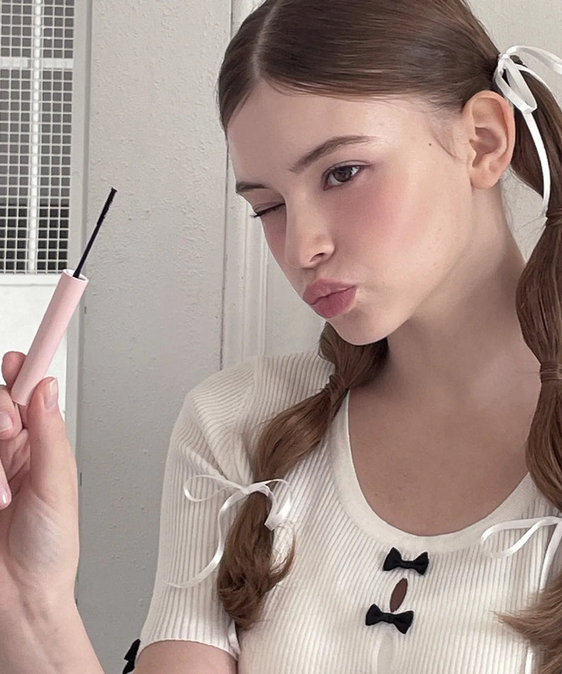 AOU - All Day Mascara de AOU Cosmetics, femme appliquant le mascara au maquillage près d’un mur clair, image produit