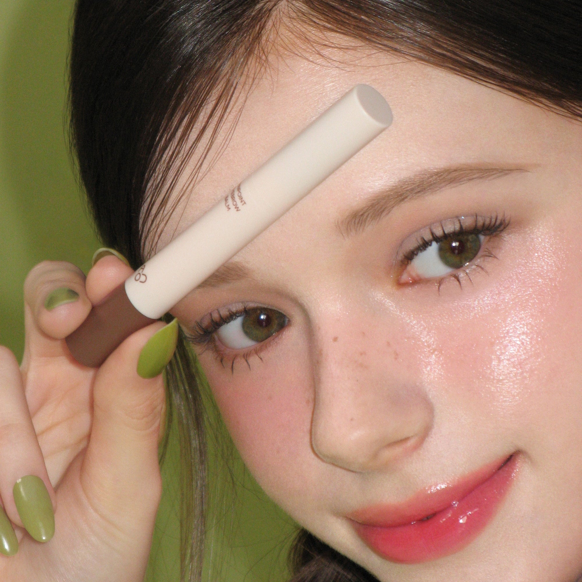 AOU Pinpoint Eyebrow Balm, tube beige sur une femme, application sur les sourcils, contexte maquillage vert.