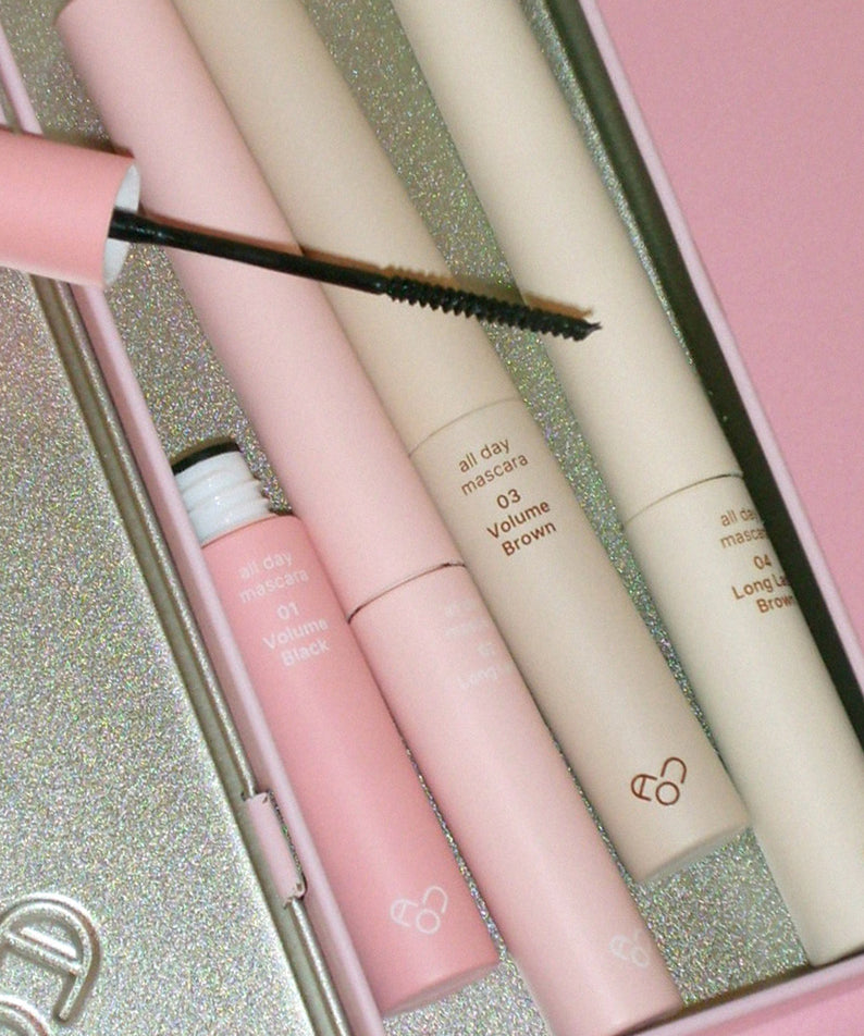 AOU - All Day Mascara de AOU COSMETICS, brosses sur coffret maquillage; contexte paillettes dorées, gros plan.
