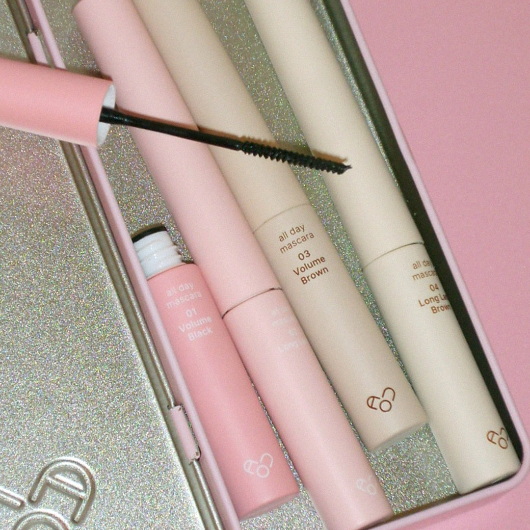 AOU - All Day Mascara de AOU COSMETICS, brosses sur coffret maquillage; contexte paillettes dorées, gros plan.