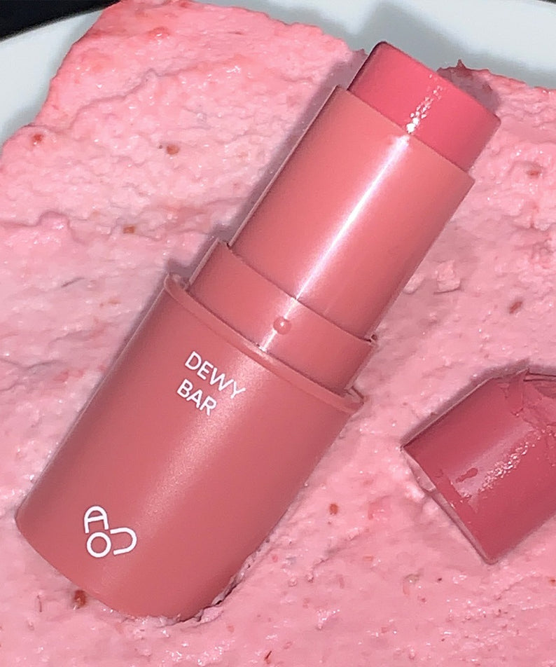 AOU Dewy Bar, marque AOU : baume hydratant rose sur fond crème texturée, capsule et étui brillants