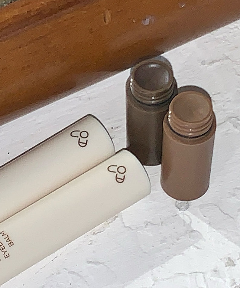 AOU Pinpoint Eyebrow Balm en deux teintes, parfait pour des sourcils définis. Cosmétique élégant sur surface marbre.