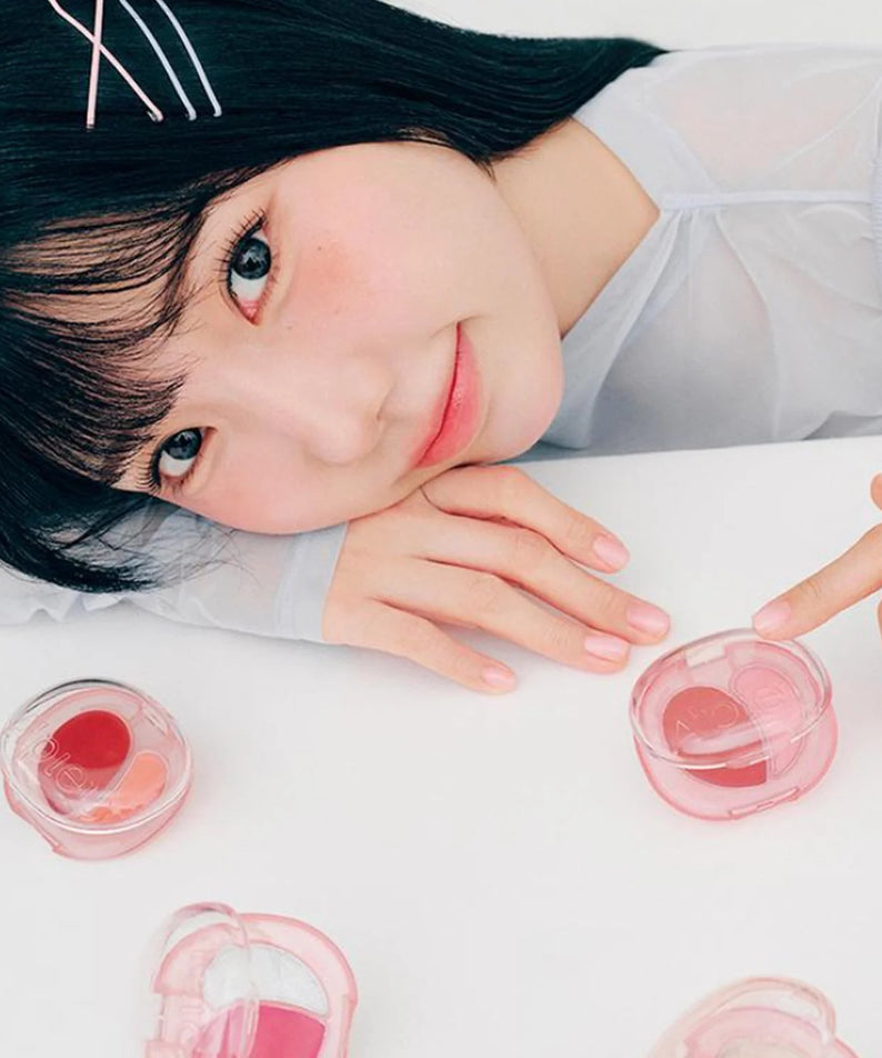 A'PIEU Juicy Pang Mood Blending Lip & Cheek Balm : femme souriante avec pots cosmétiques, ambiance beauté sur table blanche