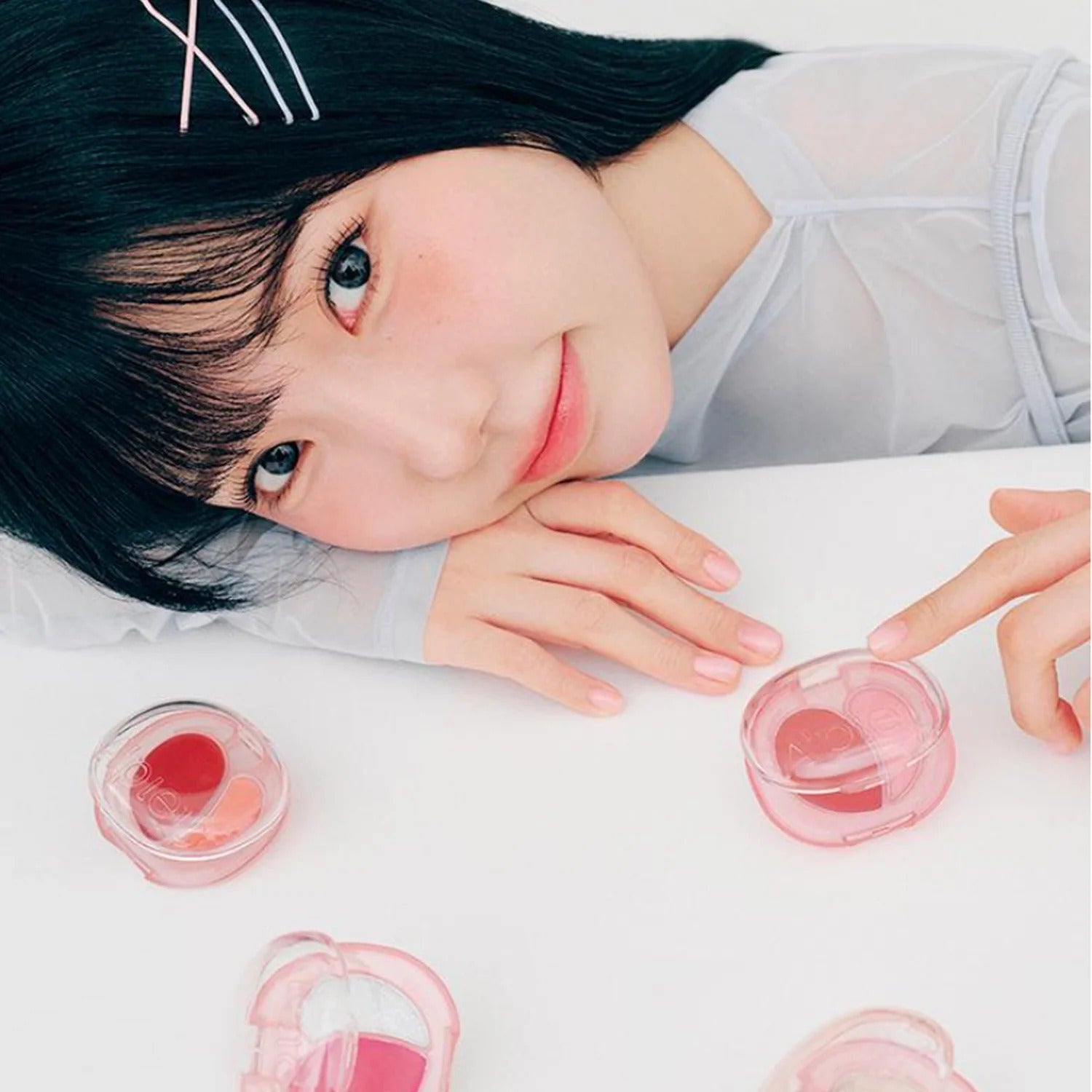 A'PIEU Juicy Pang Mood Blending Lip & Cheek Balm : femme souriante avec pots cosmétiques, ambiance beauté sur table blanche