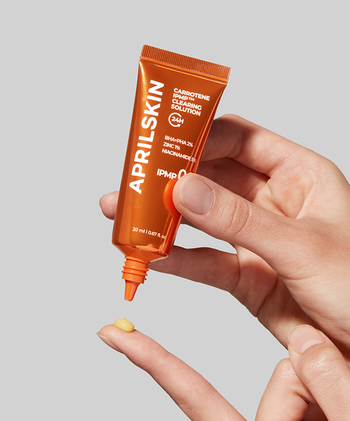Décris l'image en français : APRILSKIN Carrotene IPMP Clearing Solution d’APRILSKIN, tube orange sur main tenant un échantillon jaune.