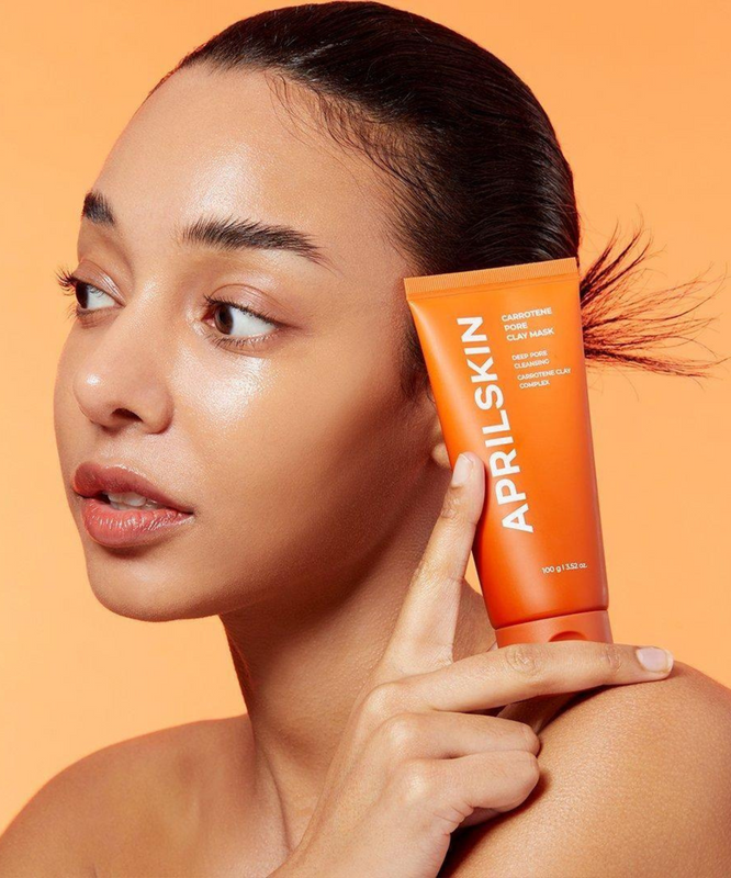 APRILSKIN Carrotene Pore Clay Mask, femme lissant le produit sur peau, packaging visible sur fond orange, APRILSKIN