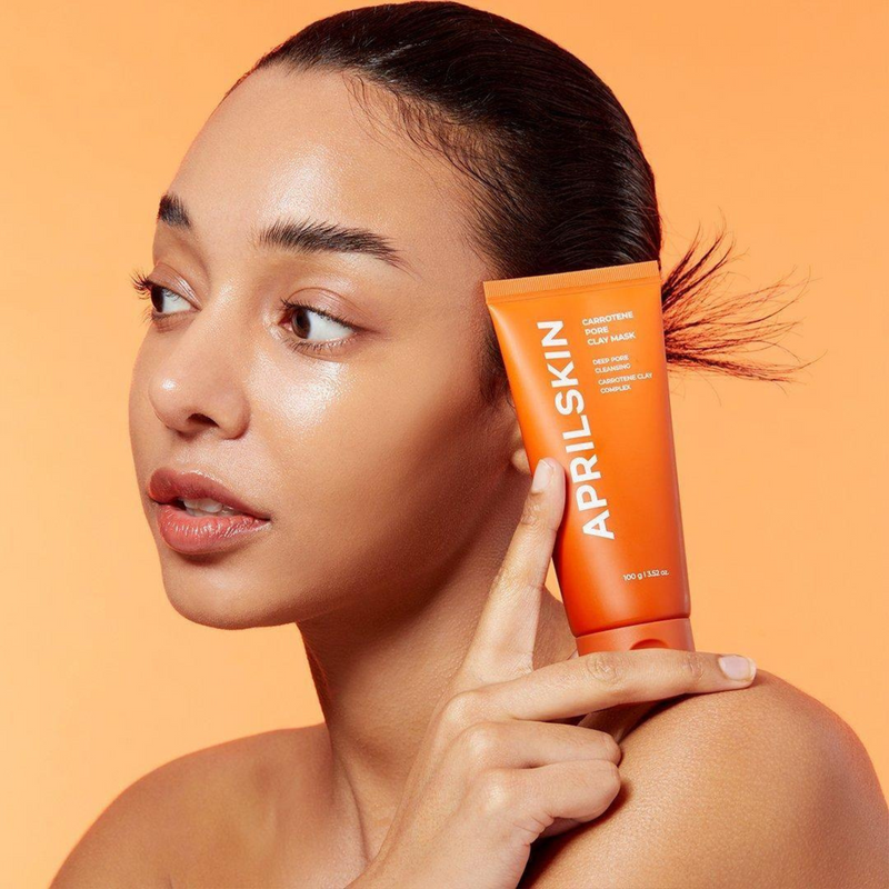 APRILSKIN Carrotene Pore Clay Mask, femme lissant le produit sur peau, packaging visible sur fond orange, APRILSKIN