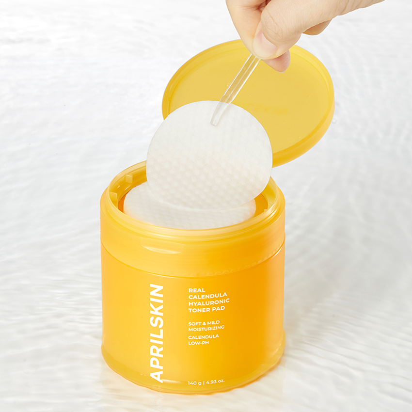 APRILSKIN Calendula Hyaluronic Toner Pad : main retire un pad du pot jaune, marque sur l’étiquette, contexte soin visage