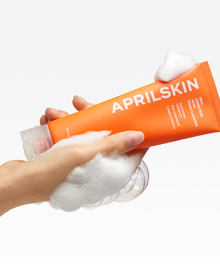 APRILSKIN Real Carrotene Acne Foam Cleanser 120ml, tube orange brandé sur main et mousse, fond blanc.